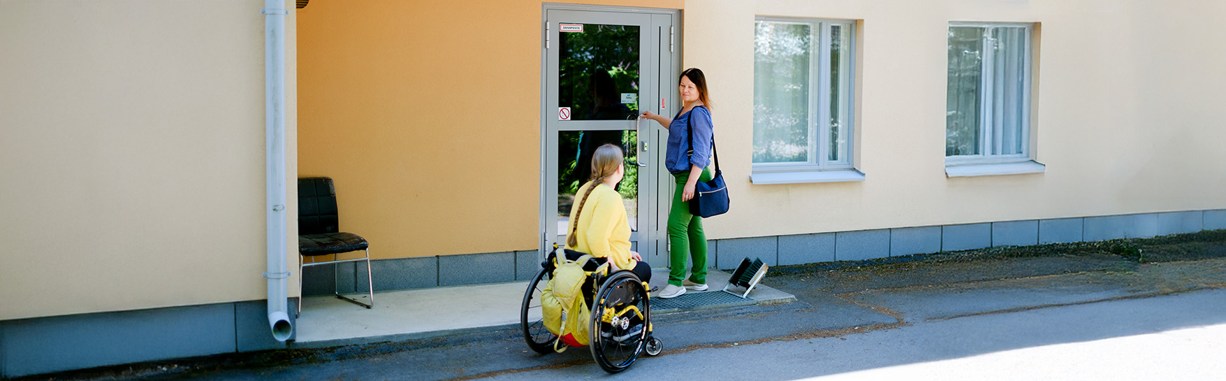 A woman approaches the door of the house in a wheelchair, where another woman is standing.