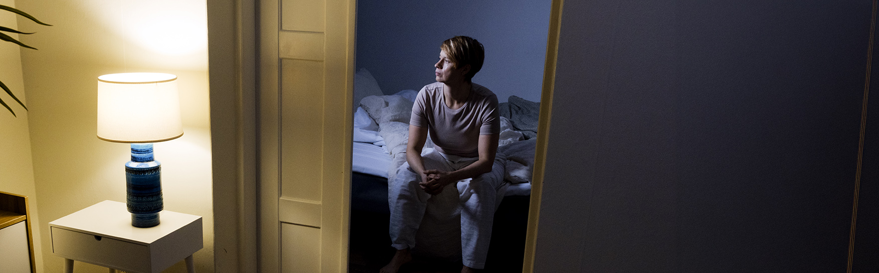 Henkilö istuu sängyllä hämärässä huoneessa. Pöytävalaisin valaisee tilaa.