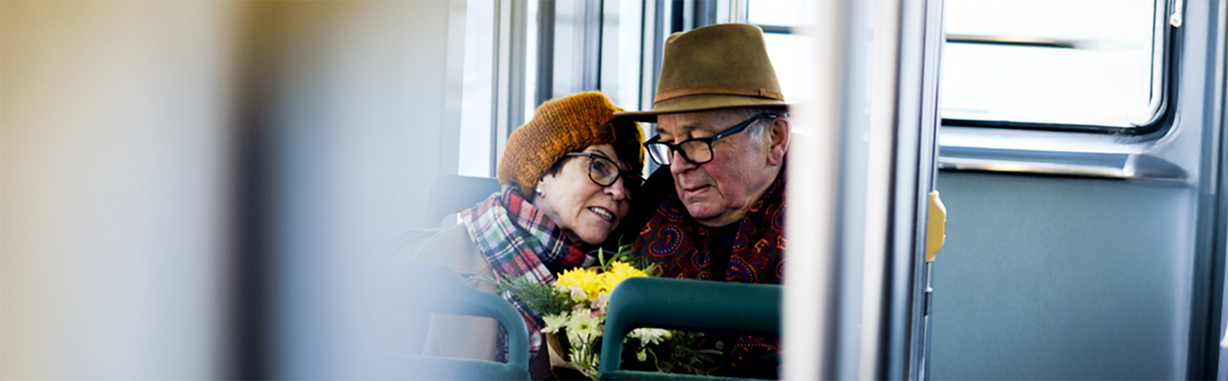 Iäkkäät nainen ja mies istuvat bussissa kukkakimppu sylissä.