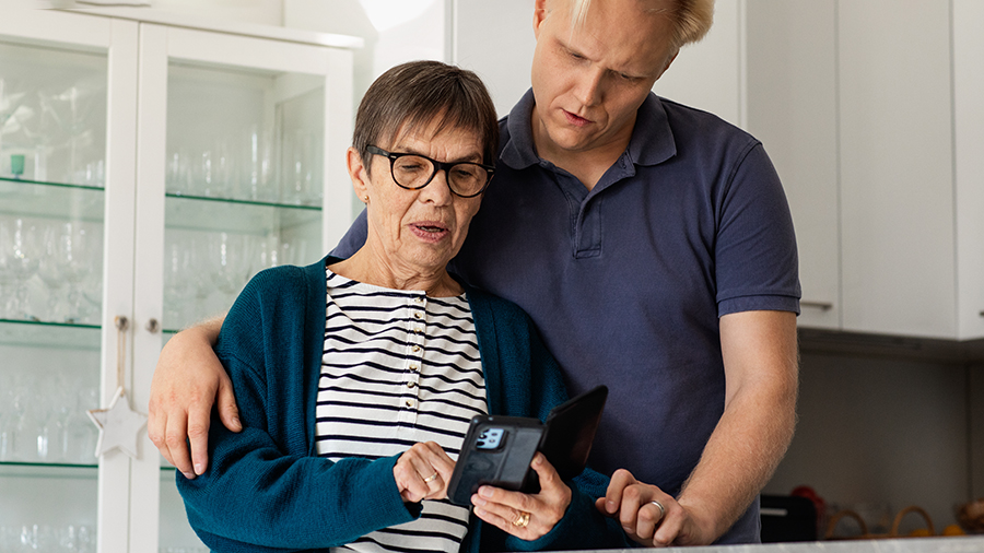 En äldre kvinna och en yngre man tittar tillsammans på sina mobiltelefoner. Mannen har sin arm runt kvinnan.