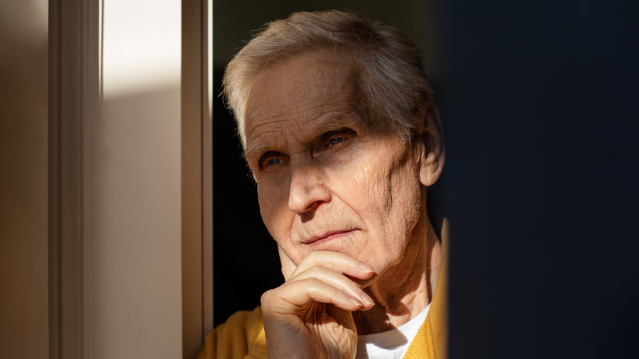 En gammal man tittar ut i fjärran, lutad mot fönsterrutan, eftertänksam.